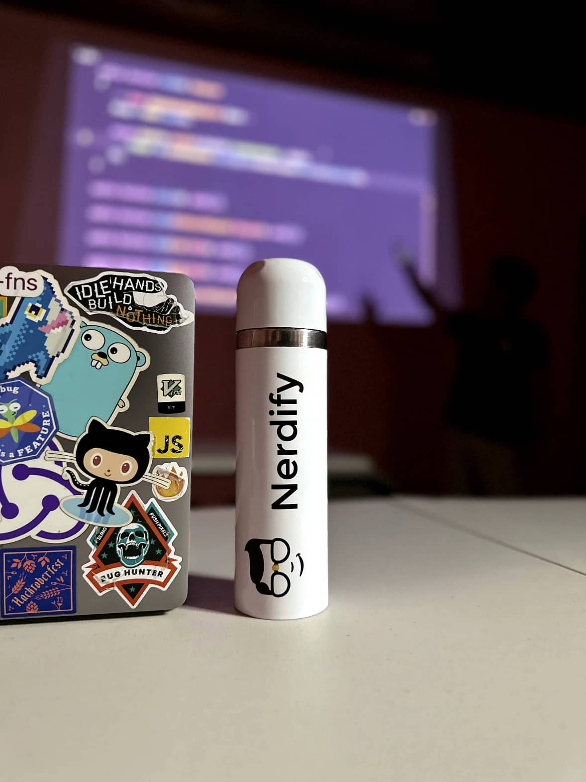 Laptop and thermos with the Nerdify logo on a table, while a developer explains code in the background